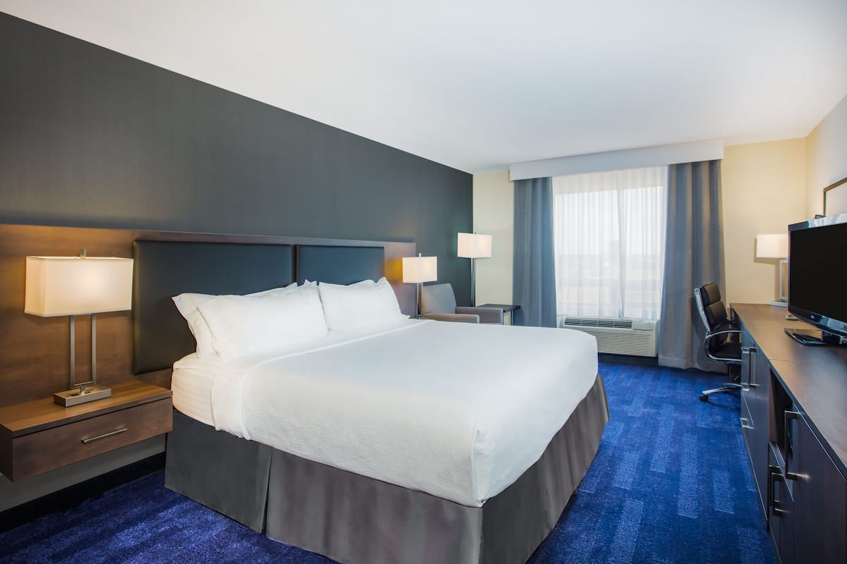 Habitación de hotel con una cama grande, dos mesitas de noche con lámparas, un escritorio con silla, un televisor y una ventana con cortinas.