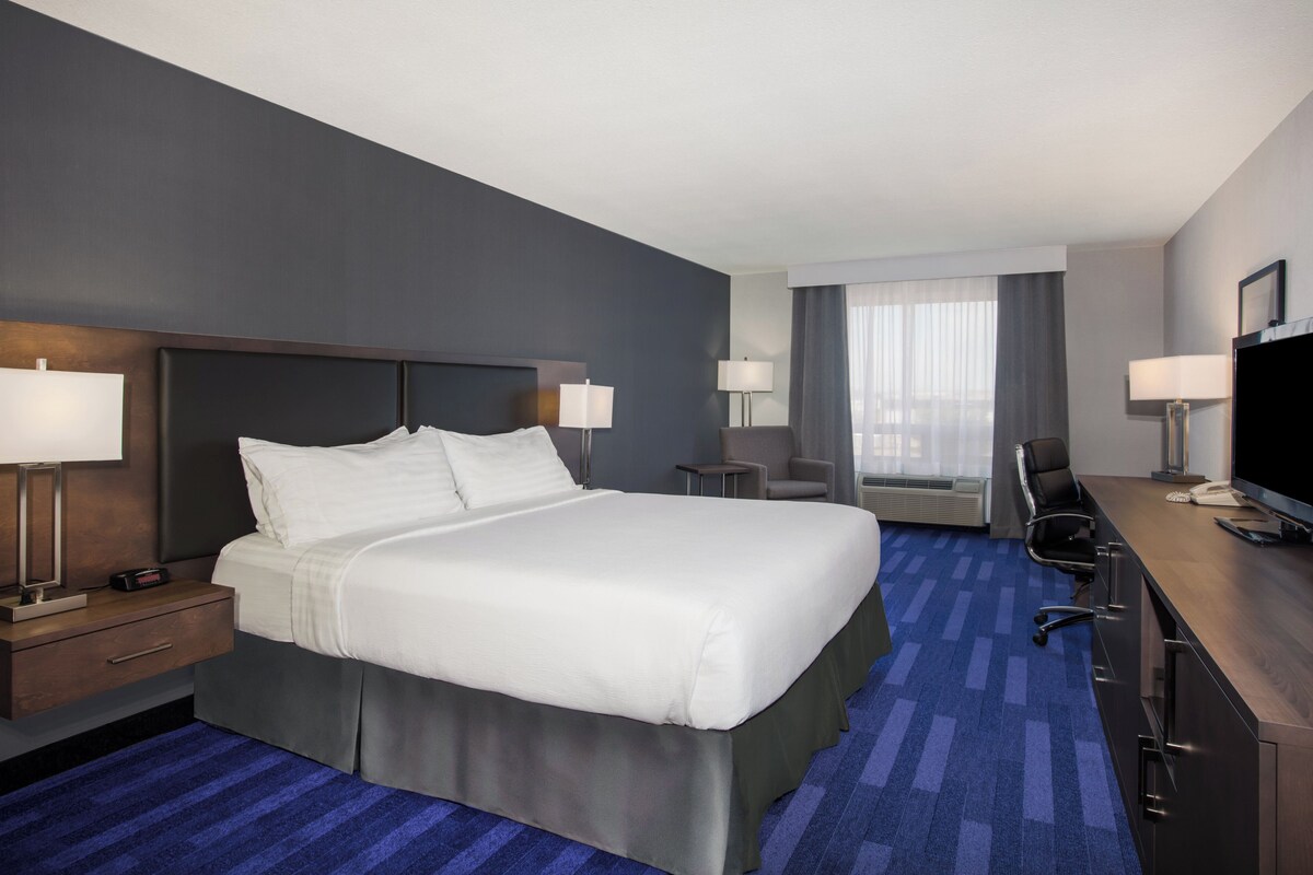 A hotel room with a large bed, a desk with a computer, a chair, a TV, and a window with curtains.