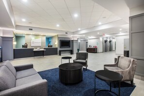 A hotel lobby with seating, a reception desk, a TV, and a staircase leading to elevators.