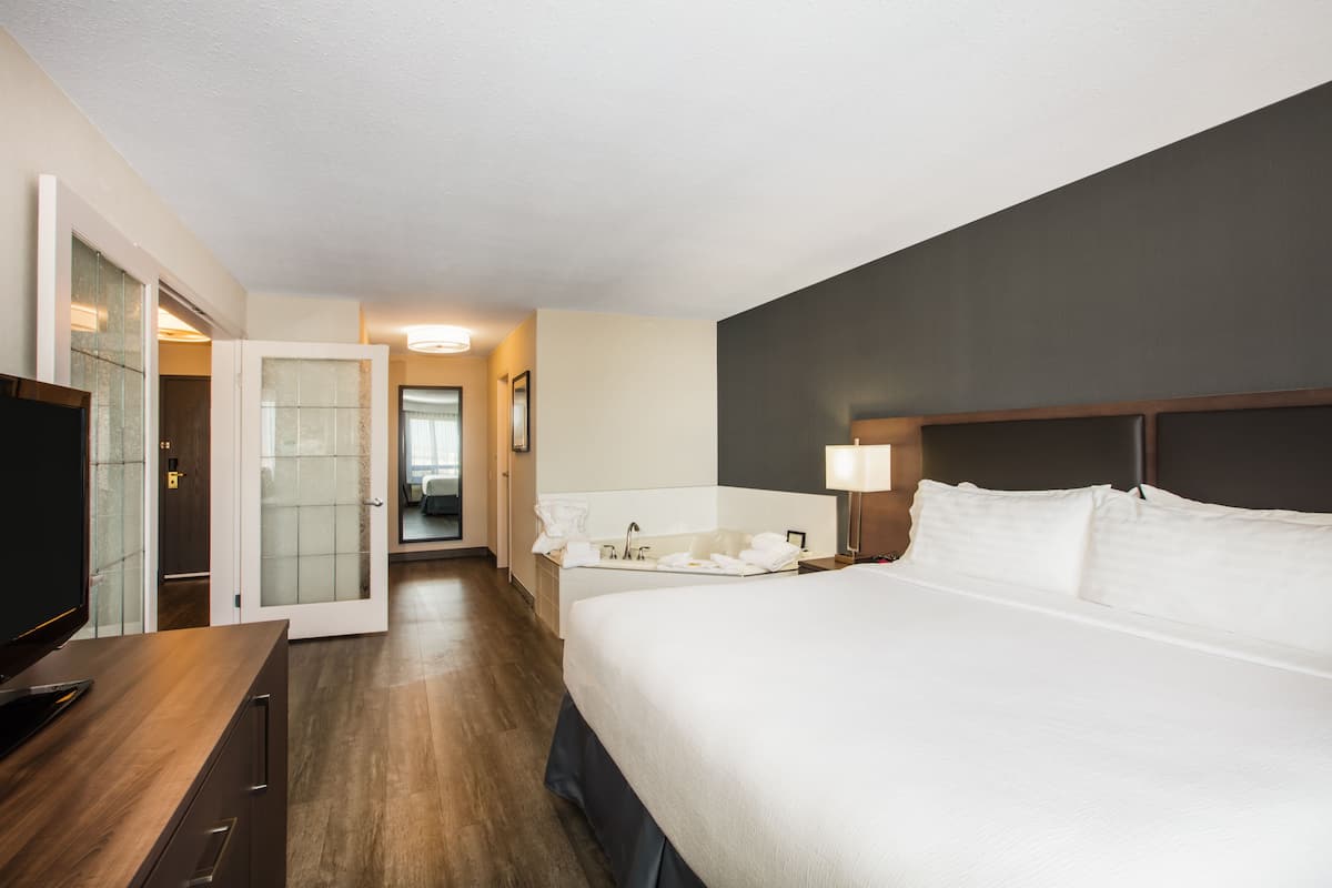 Habitación de hotel con una cama grande, un escritorio con televisor de pantalla plana, un baño con lavamanos y espejo, y una puerta que da a otra habitación.
