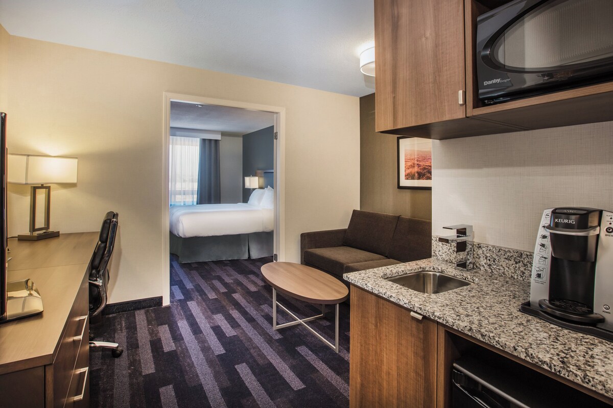 A compact hotel room with a kitchenette, a bed, a small seating area, and a view of the hallway.