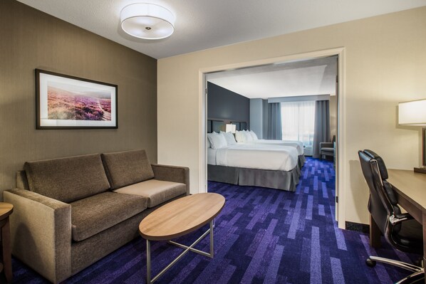 A hotel room with a sofa, a small table, a desk with a chair, a bed, and a bathroom visible through an open door.