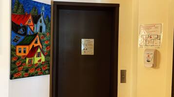 A hotel room with a dark door, a painting of colorful houses, a COVID-19 informational sign, and a hand sanitizer dispenser.