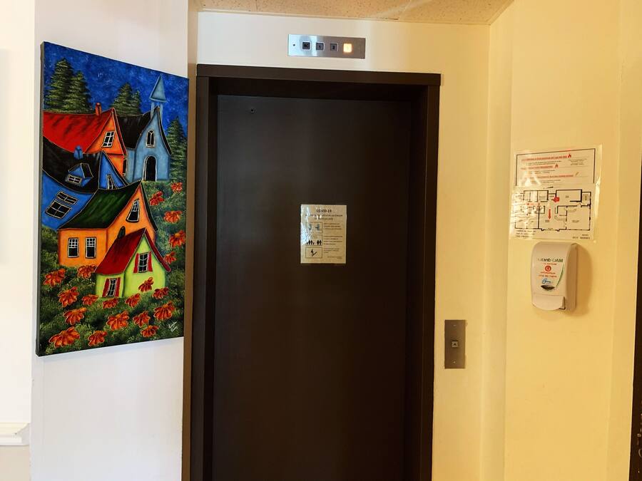 A hotel room with a dark door, a painting of colorful houses, a COVID-19 informational sign, and a hand sanitizer dispenser.