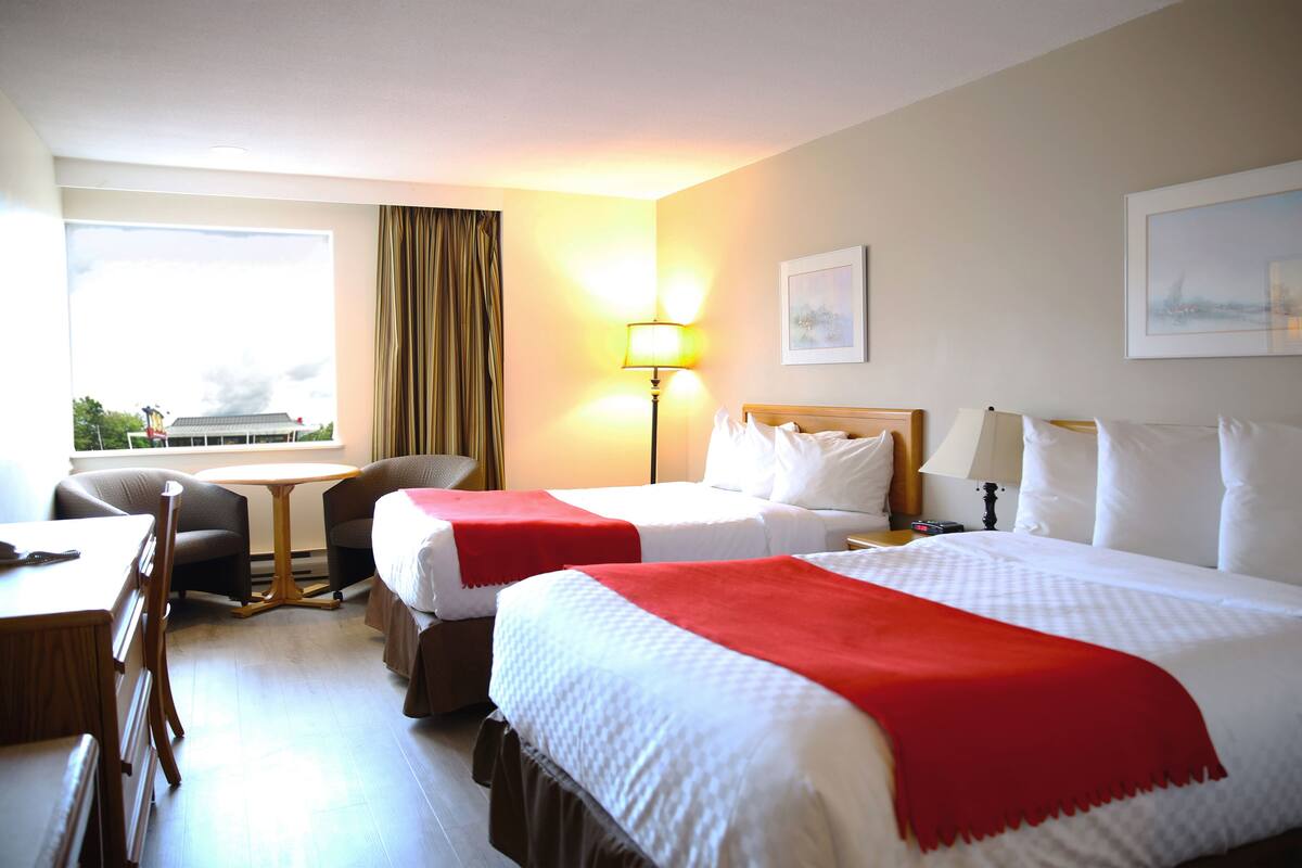A hotel room with two beds, a desk, a chair, a lamp, and a window with curtains.