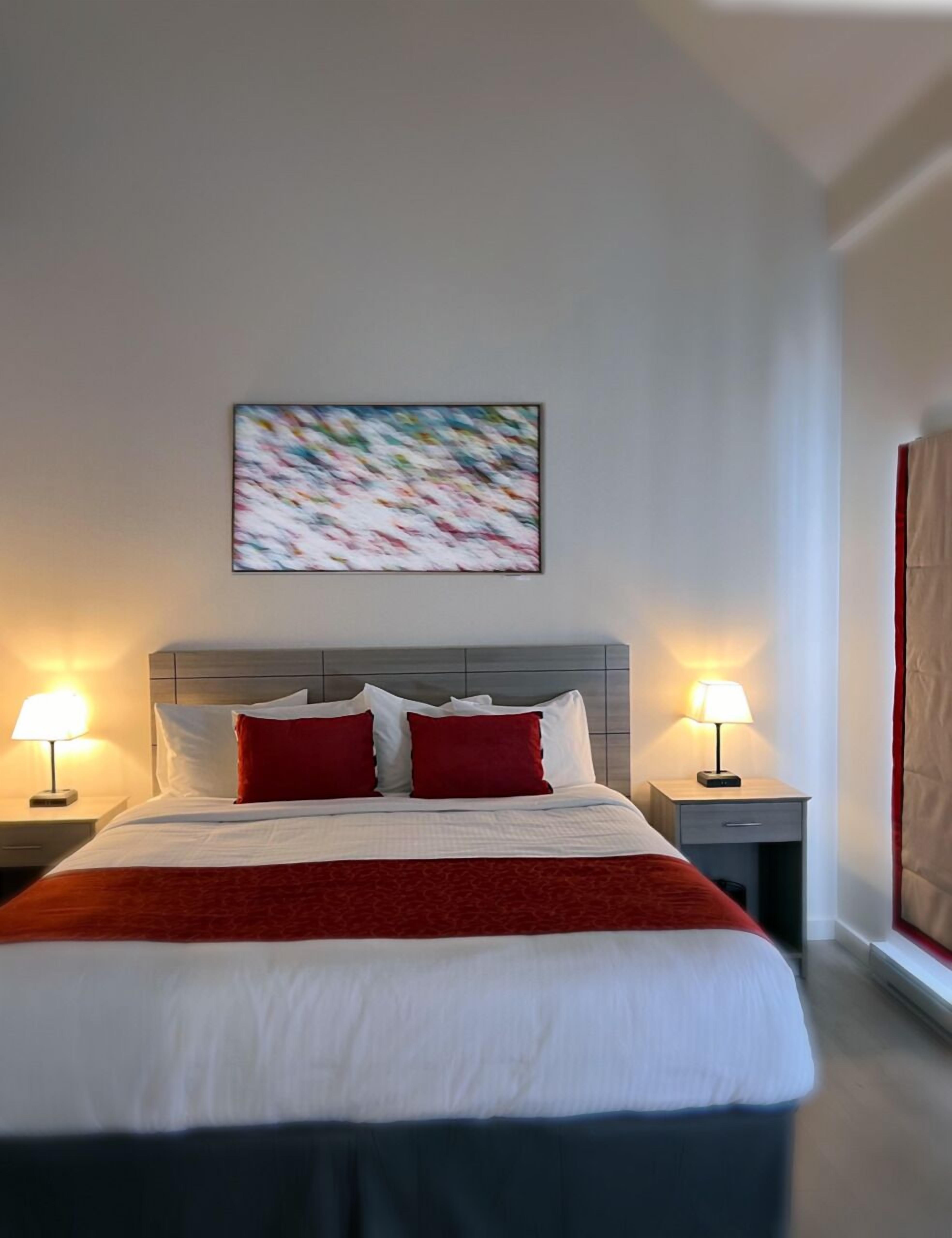 A hotel room with a bed, two bedside lamps, a red and white bedspread, a gray headboard, and a painting on the wall.