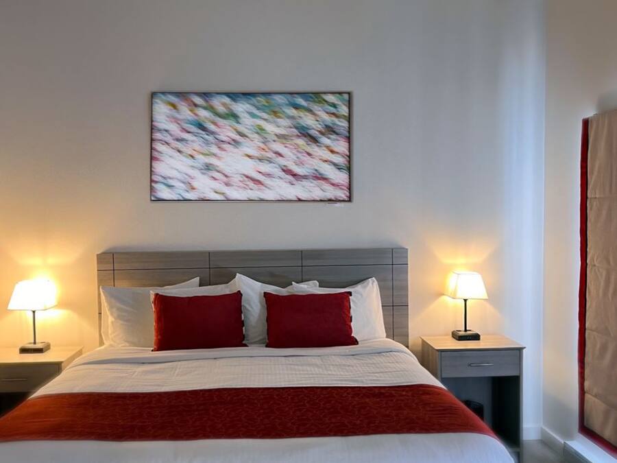 Habitación de hotel con cama, dos lámparas de noche, colcha roja y blanca, cabecero gris y un cuadro en la pared.