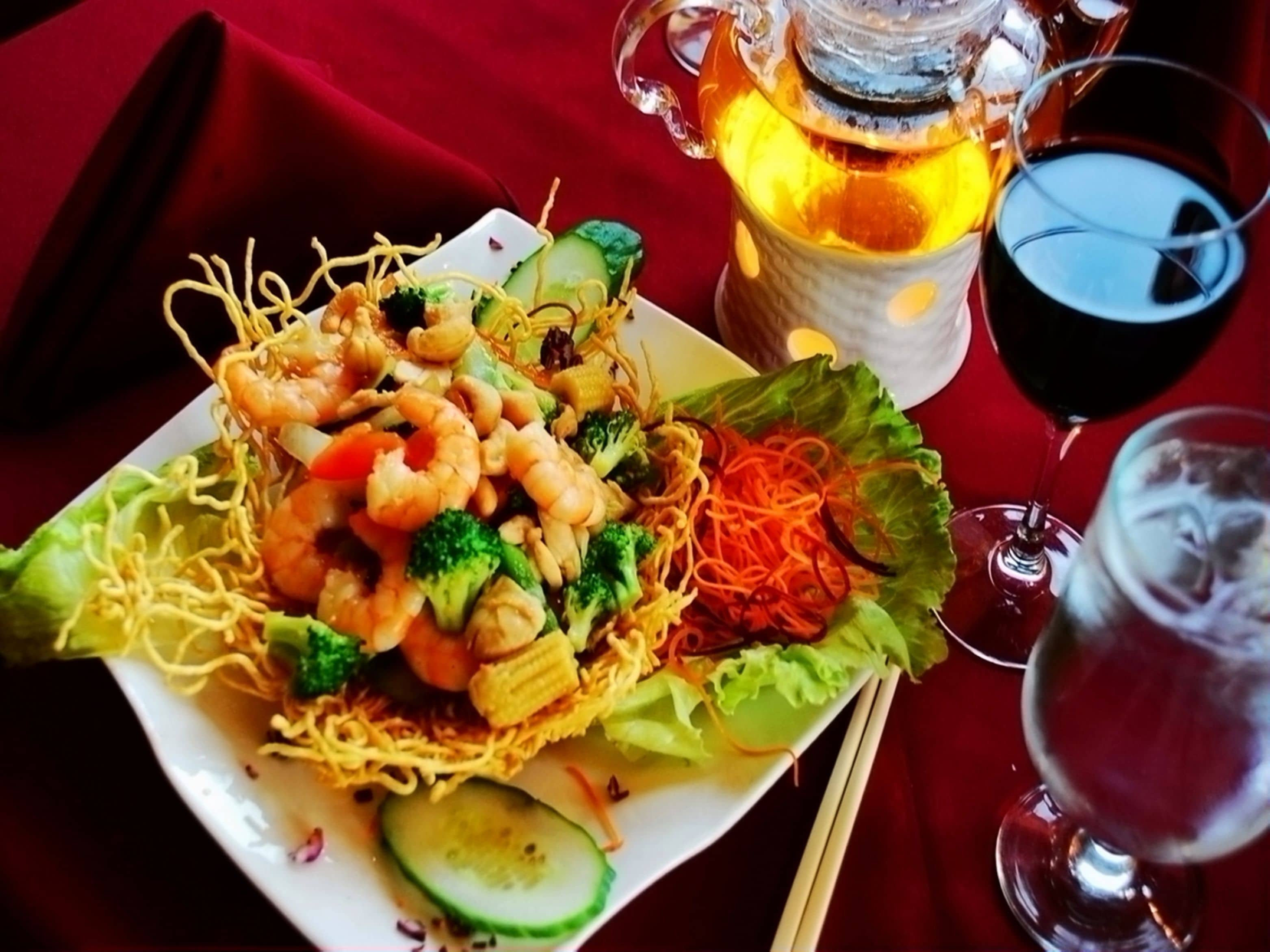 Une assiette de nouilles aux crevettes et aux légumes, une carafe de thé et un verre de vin rouge sur une table.
