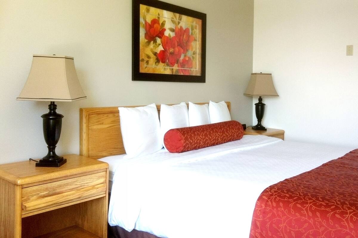 A hotel room with a wooden headboard, white bedding, a red decorative pillow, two bedside tables with lamps, and a framed floral artwork on the wall.