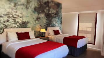 A hotel room with two beds, each with white linens and red accents, a nightstand with a lamp, and a large abstract mural on the wall.