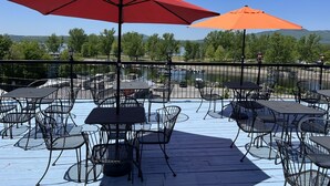 An outdoor patio with tables and chairs, red and orange umbrellas, and a view of a river and mountains. - Auberge Du Grand Lac (Magog)