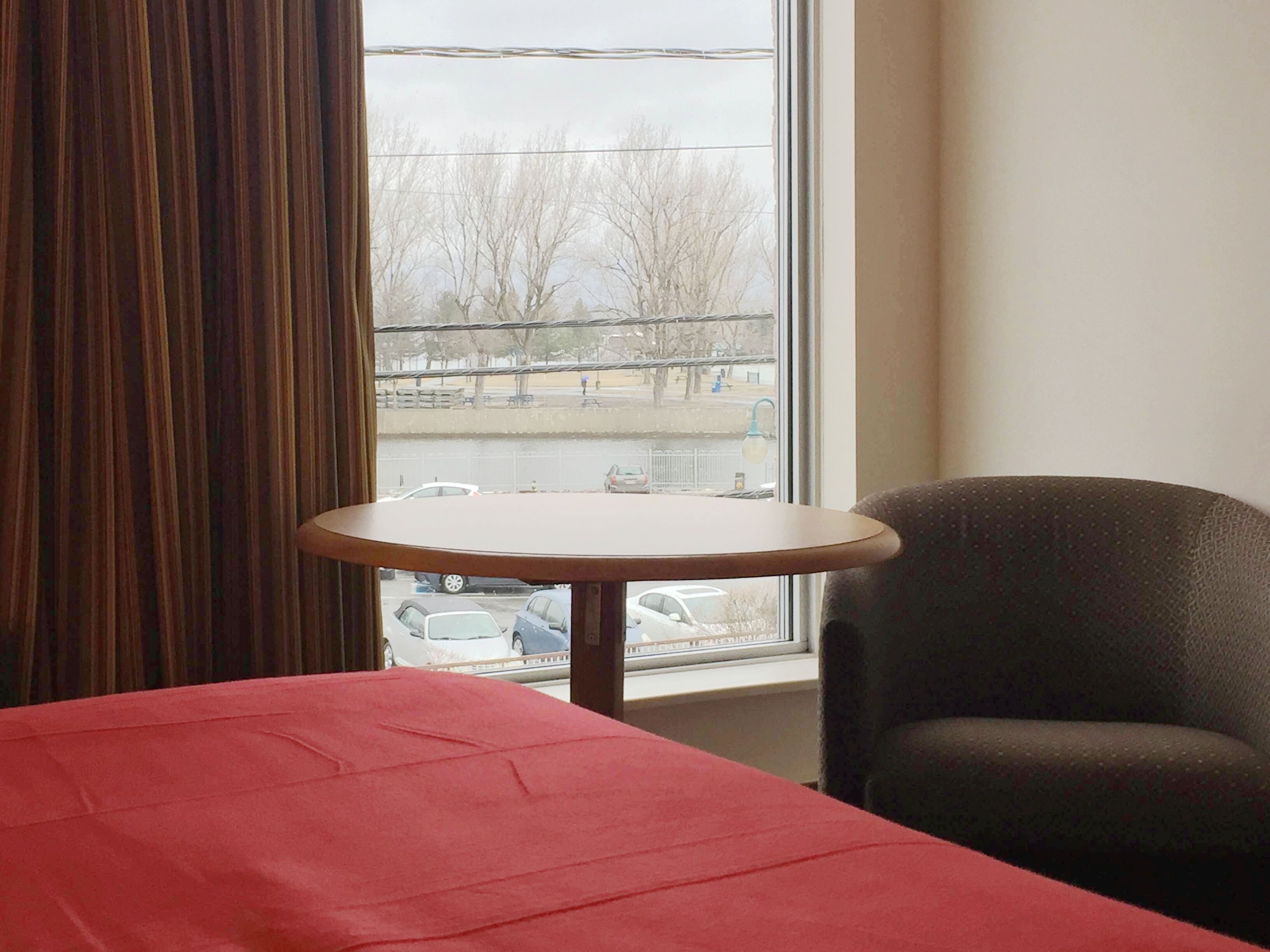 A hotel room with a bed, a small round table, a chair, and a window with a view of a parking lot and trees.
