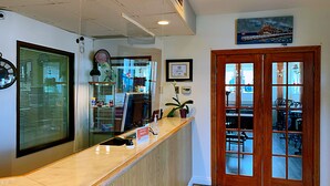 A reception area with a wooden counter, a large window with blinds, a framed certificate on the wall, and a view of the dining area through a glass door. - Auberge Du Grand Lac (Magog)