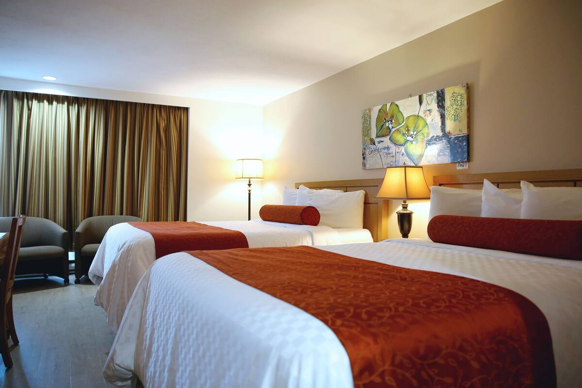 A hotel room with two beds, a desk, a chair, a lamp, and a painting on the wall.