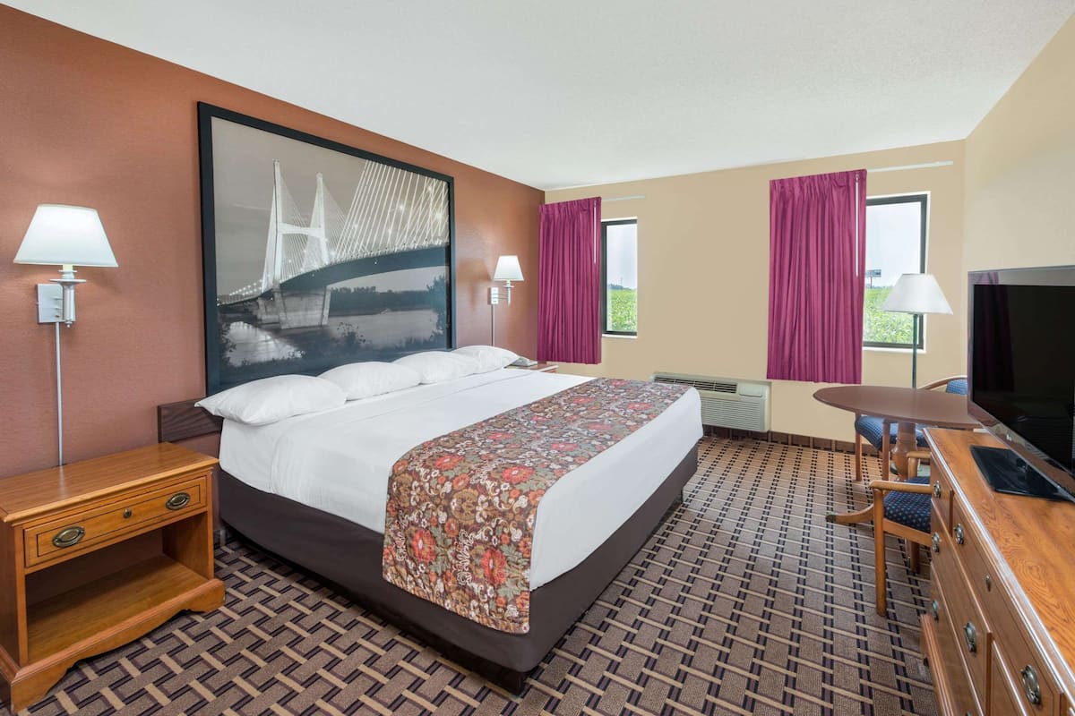 Ein Hotelzimmer mit einem großen Bett, einem Schreibtisch mit Fernseher, zwei Lampen und einem Fenster mit Vorhängen.