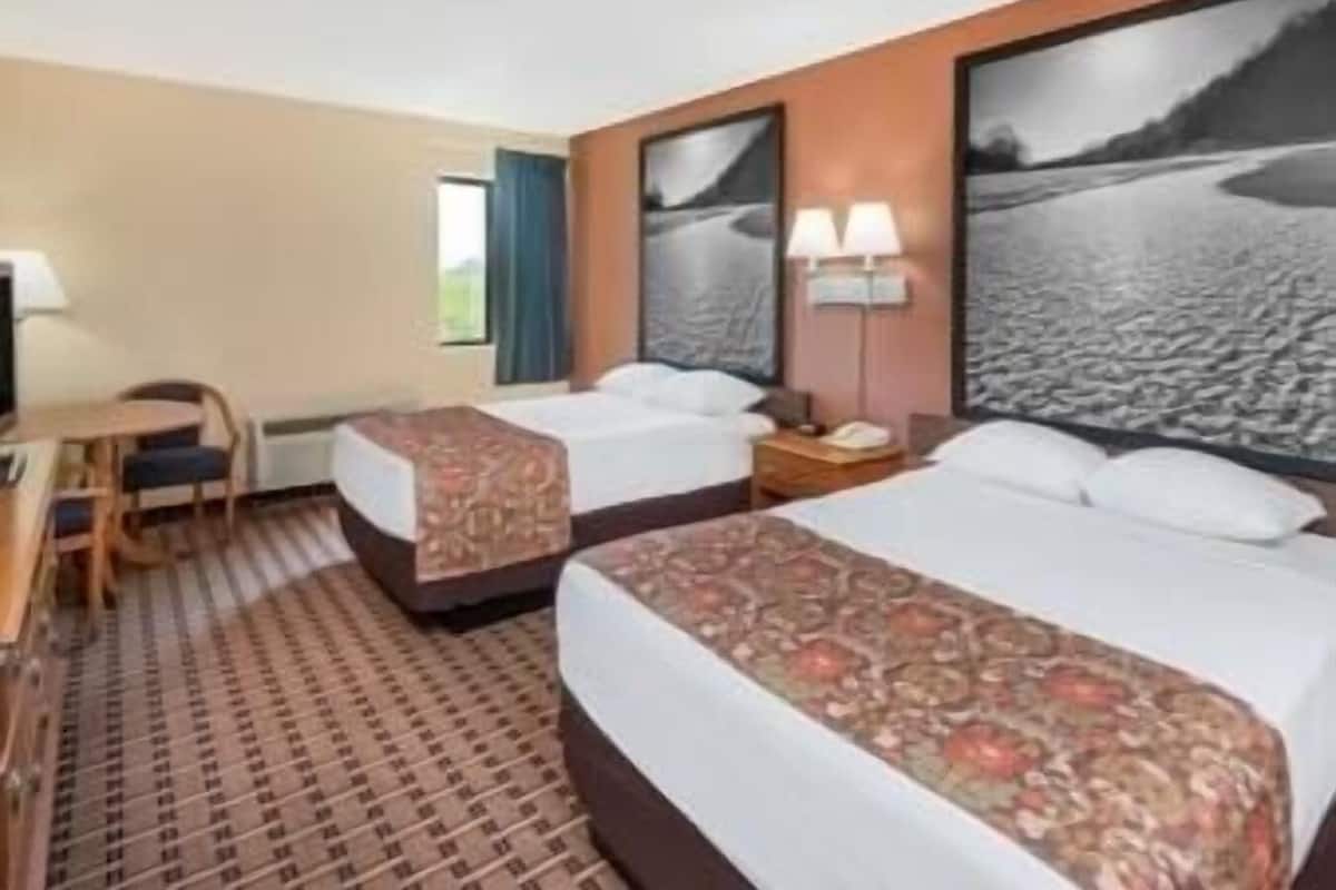 Ein Hotelzimmer mit zwei Betten, einem Schreibtisch, einem Stuhl, einer Lampe und einem großen Bild in einem Rahmen an der Wand.