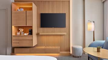 A modern hotel room with a wooden panel wall, a flat-screen TV, a sofa, a small round table, and a seating area with a bench.