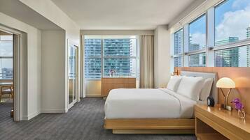 A hotel room with a large bed, a bedside table with a lamp, a window with a city view, and a door leading to a balcony.