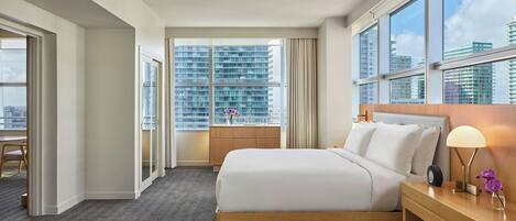 A hotel room with a large bed, a bedside table with a lamp, a window with a city view, and a door leading to a balcony.