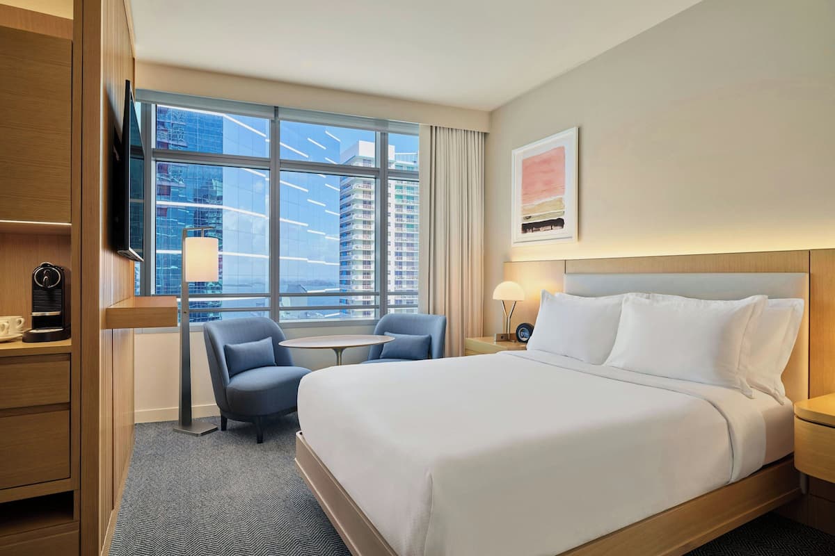 Habitación de hotel con cama, escritorio, silla, mesita, televisor y ventana con vista a la ciudad.