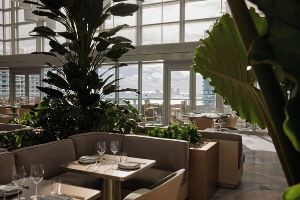 A modern restaurant with high ceilings, large windows, and indoor plants. Tables are set with wine glasses and plates. - Brickell Arch, a Luxury Collection Hotel, Miami (Miami)