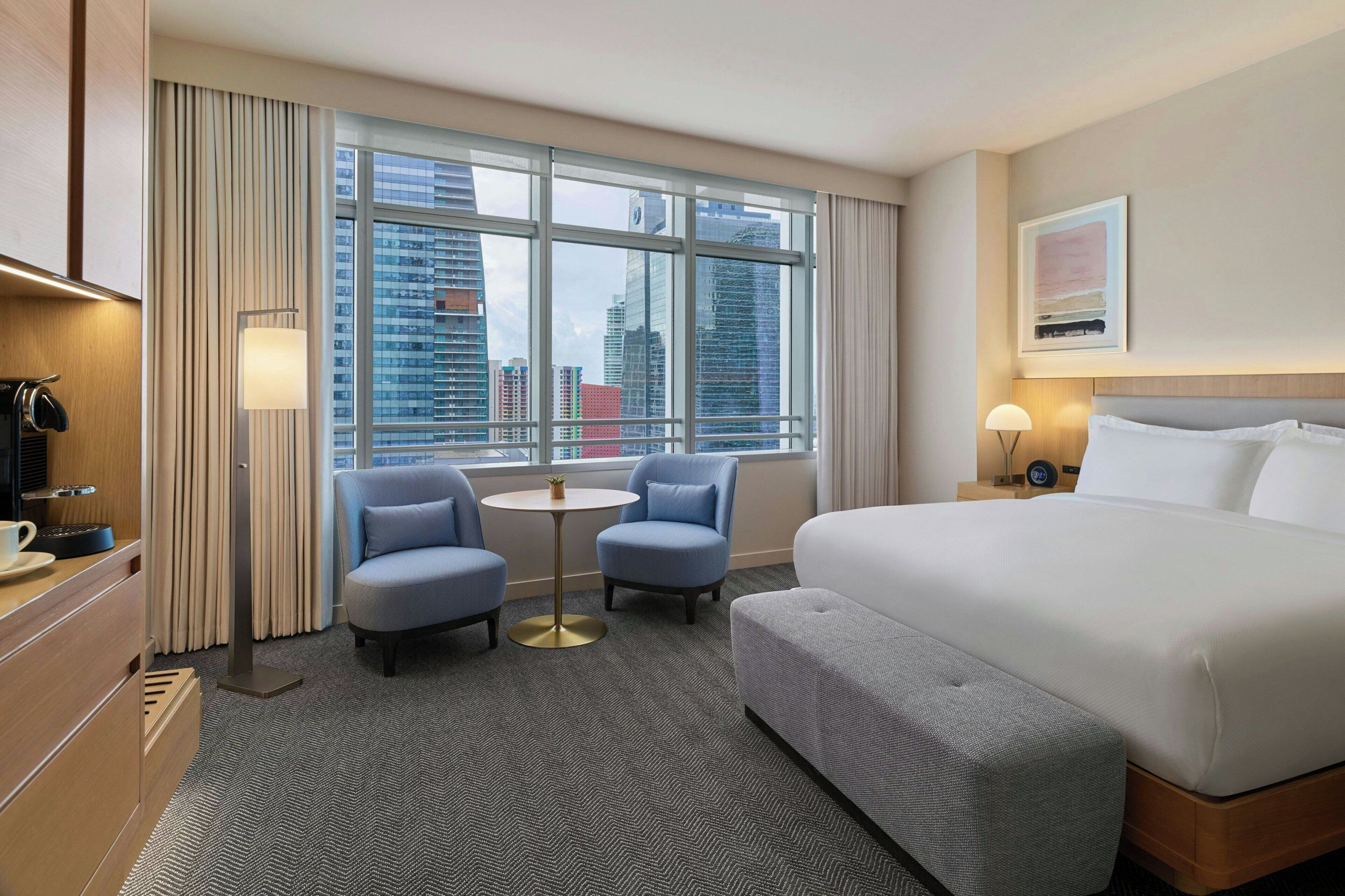 A hotel room with a large bed, two armchairs, a small table, and a view of the cityscape through large windows.