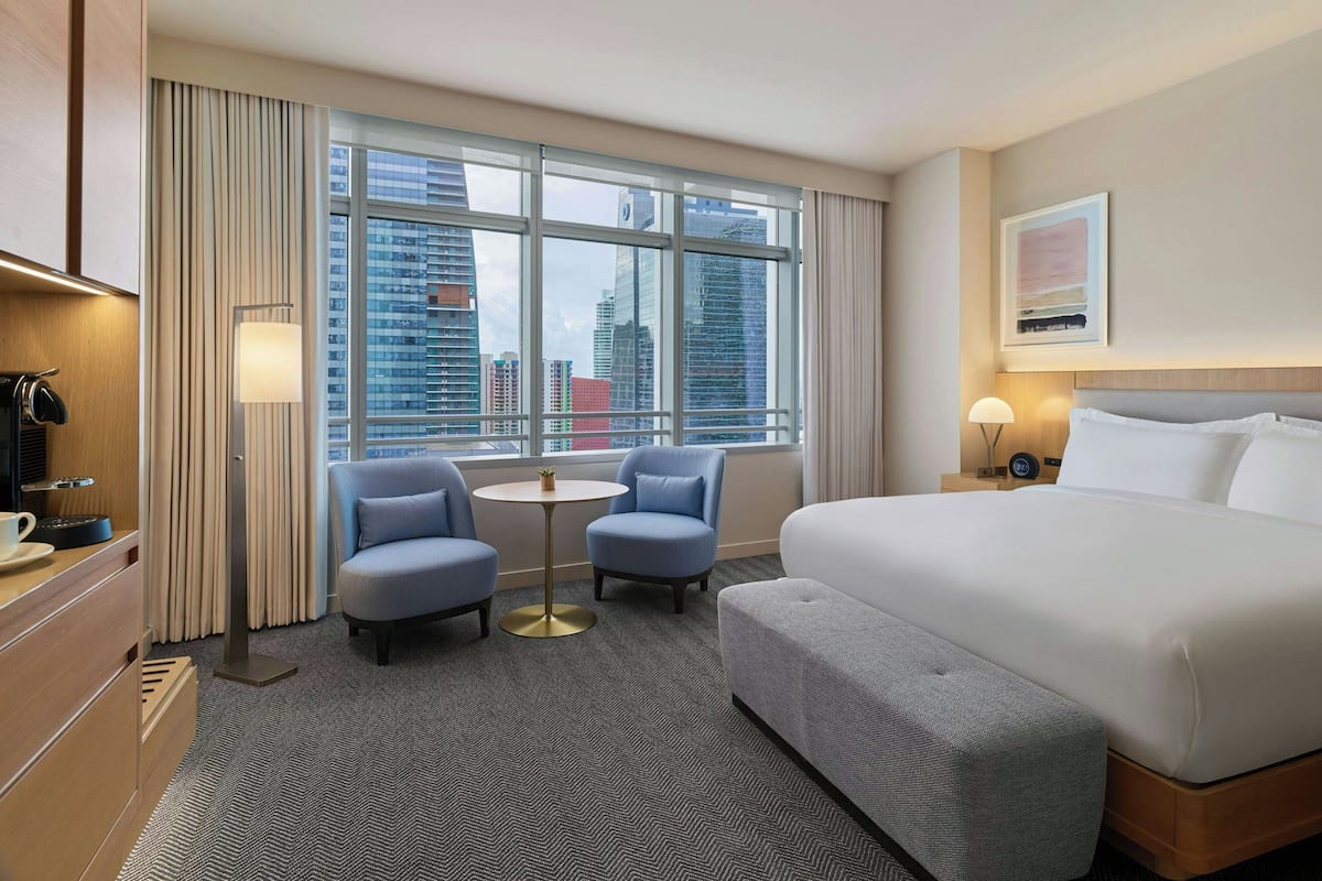 Habitación de hotel con una cama grande, dos sillones, una mesita y vistas a la ciudad a través de amplios ventanales.