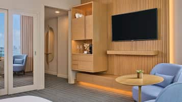 Habitación de hotel moderna con un televisor grande montado en una pared de madera, una zona de estar con una mesa redonda y vistas a la ciudad a través de amplias ventanas.