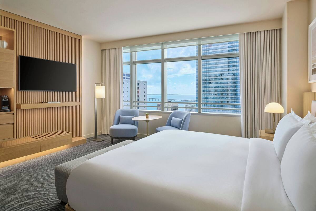 Habitación de hotel con una cama grande, dos sillones, una mesita, televisor y vistas a la ciudad.