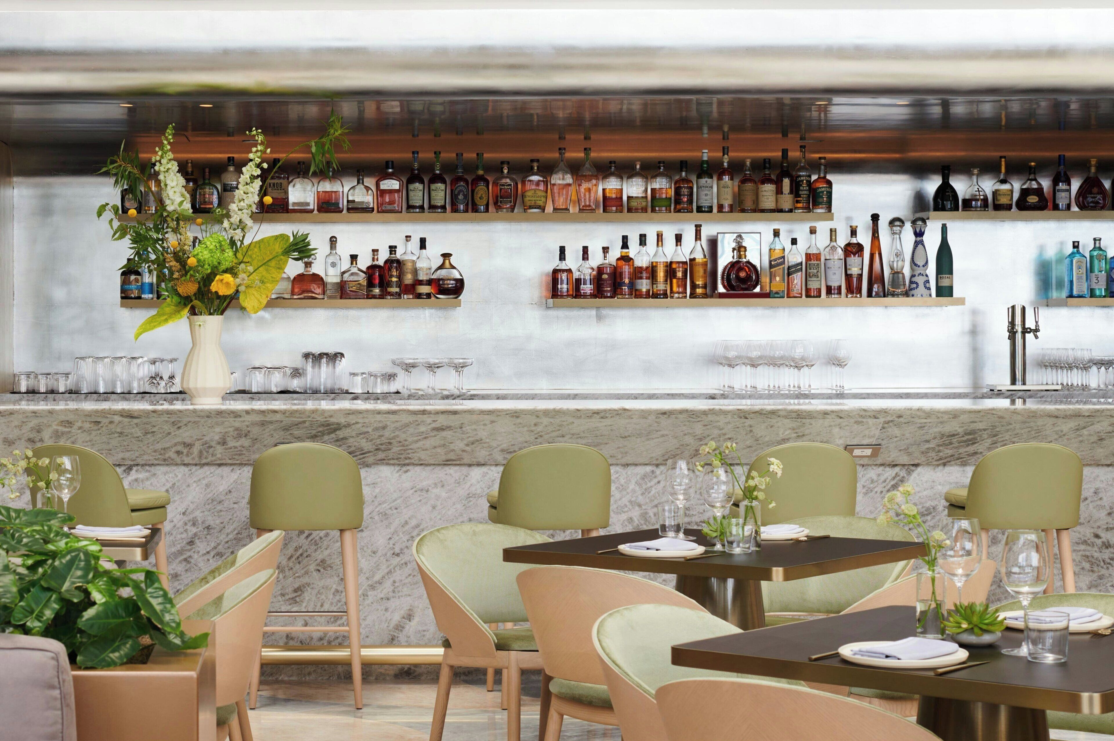 A restaurant with a bar area displaying various liquor bottles, green upholstered chairs, marble countertops, and a vase with flowers.