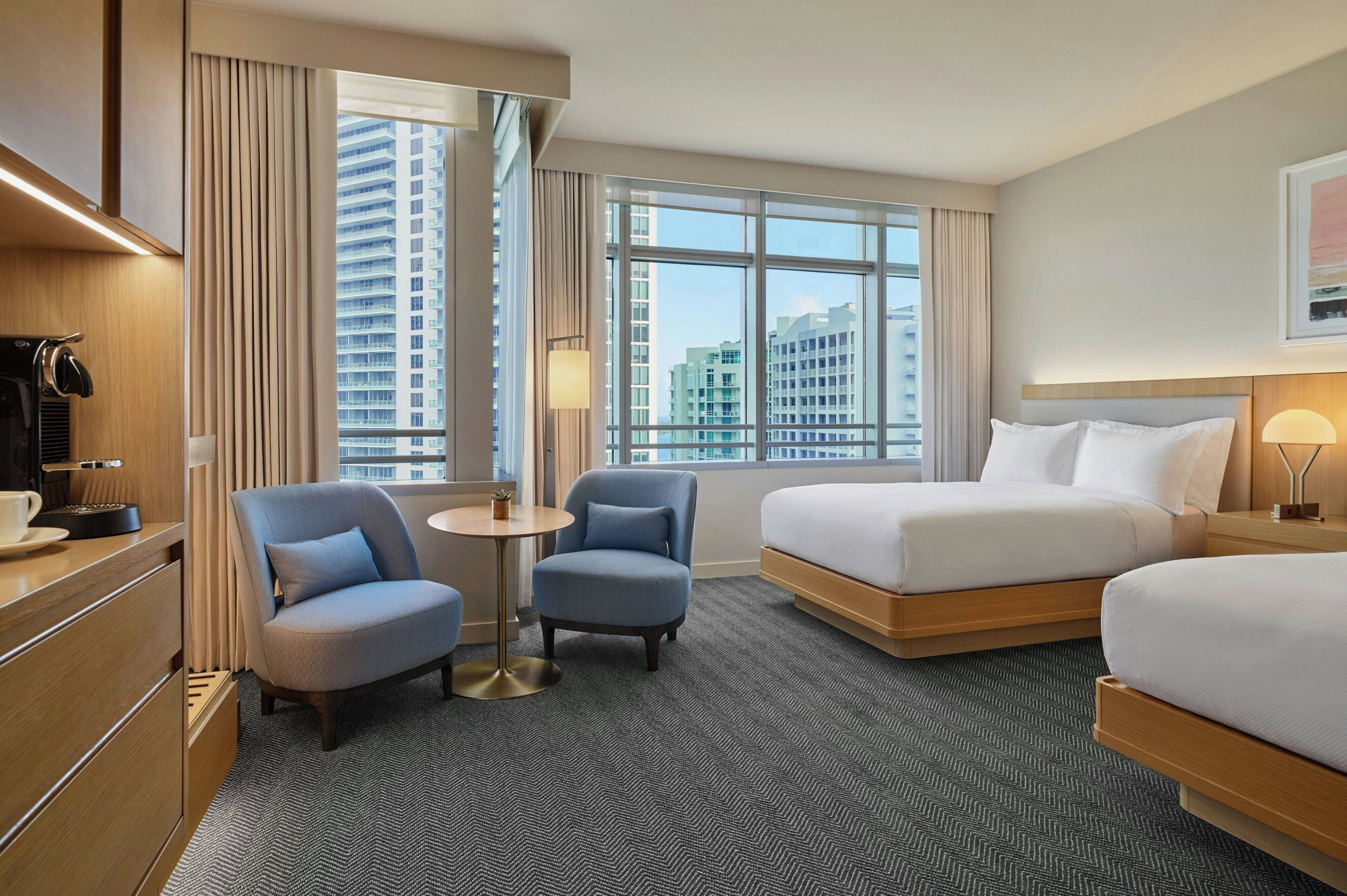 A hotel room with a bed, a chair, a small table, and a view of tall buildings.