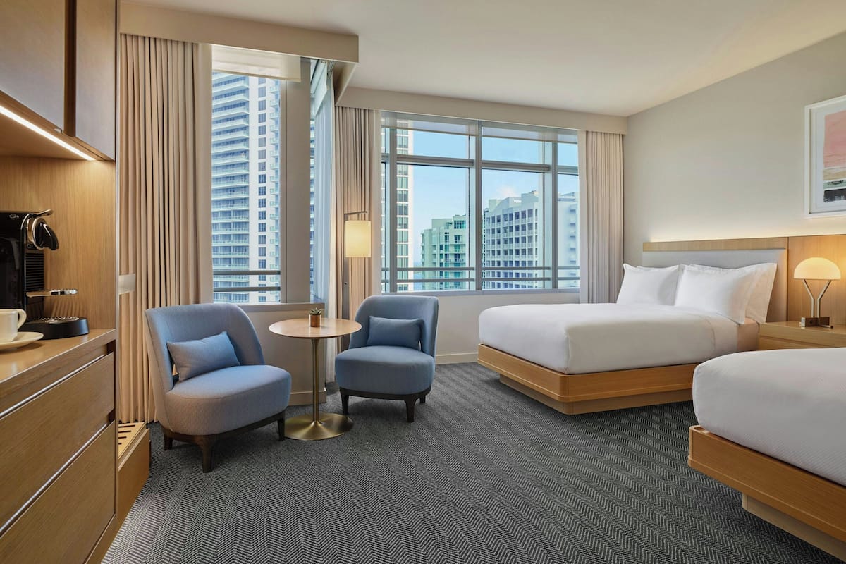 A hotel room with a bed, a chair, a small table, and a view of tall buildings.