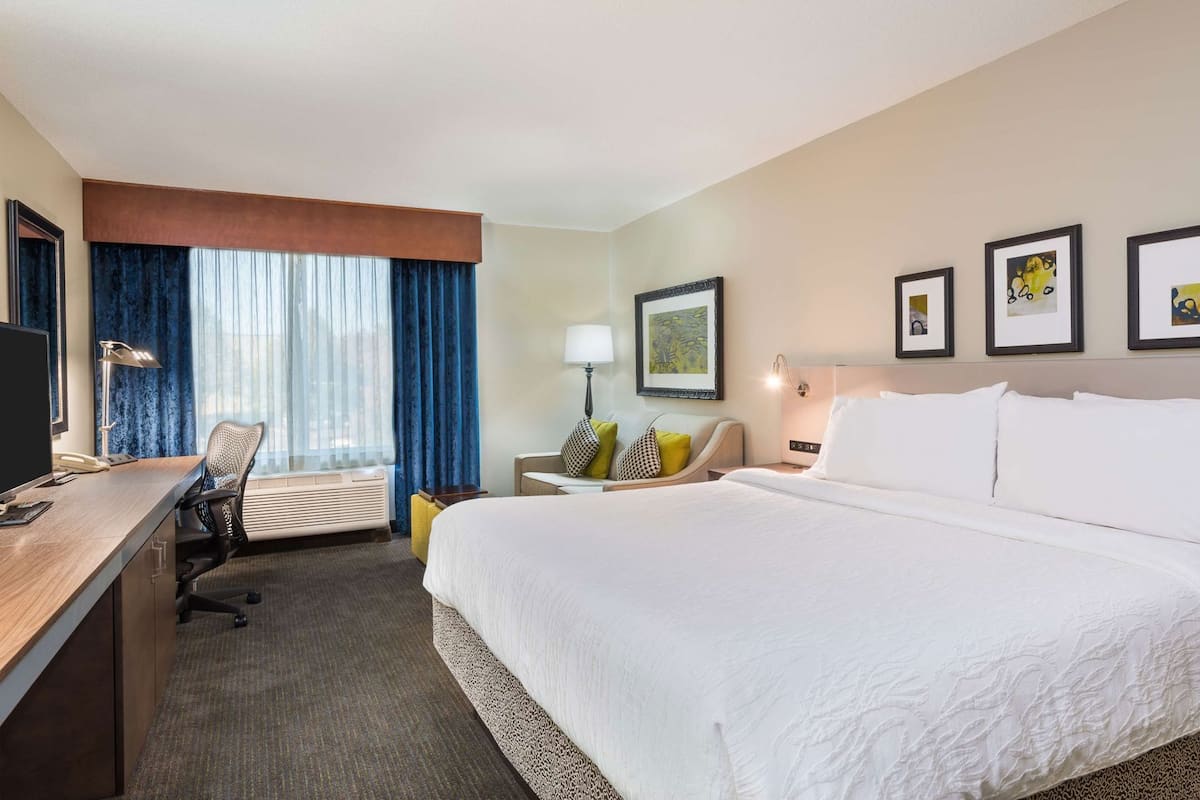 Habitación de hotel con una cama grande, un escritorio con computadora, una silla y una ventana con cortinas.