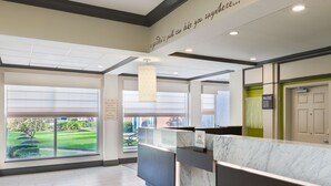 A hotel reception area with a marble counter, large windows offering a view of a lawn and building, and a ceiling with recessed lighting. - Hilton Garden Inn Westbury (Westbury)