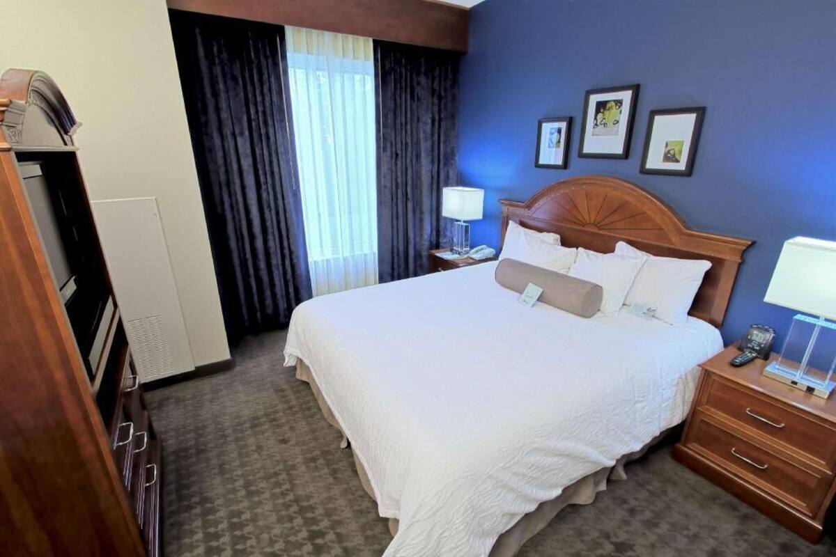 Una habitación de hotel con cama, mesita de noche, escritorio, silla, ventana con cortinas y pared decorada con cuadros.