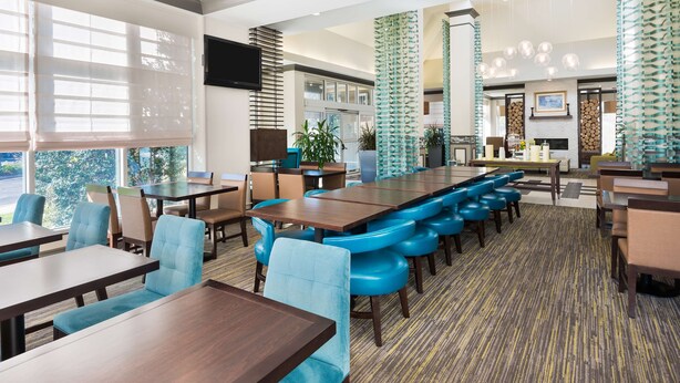 A modern hotel lobby with a long communal table, blue cushioned seating, and a patterned carpet. - Hilton Garden Inn Westbury (Westbury)