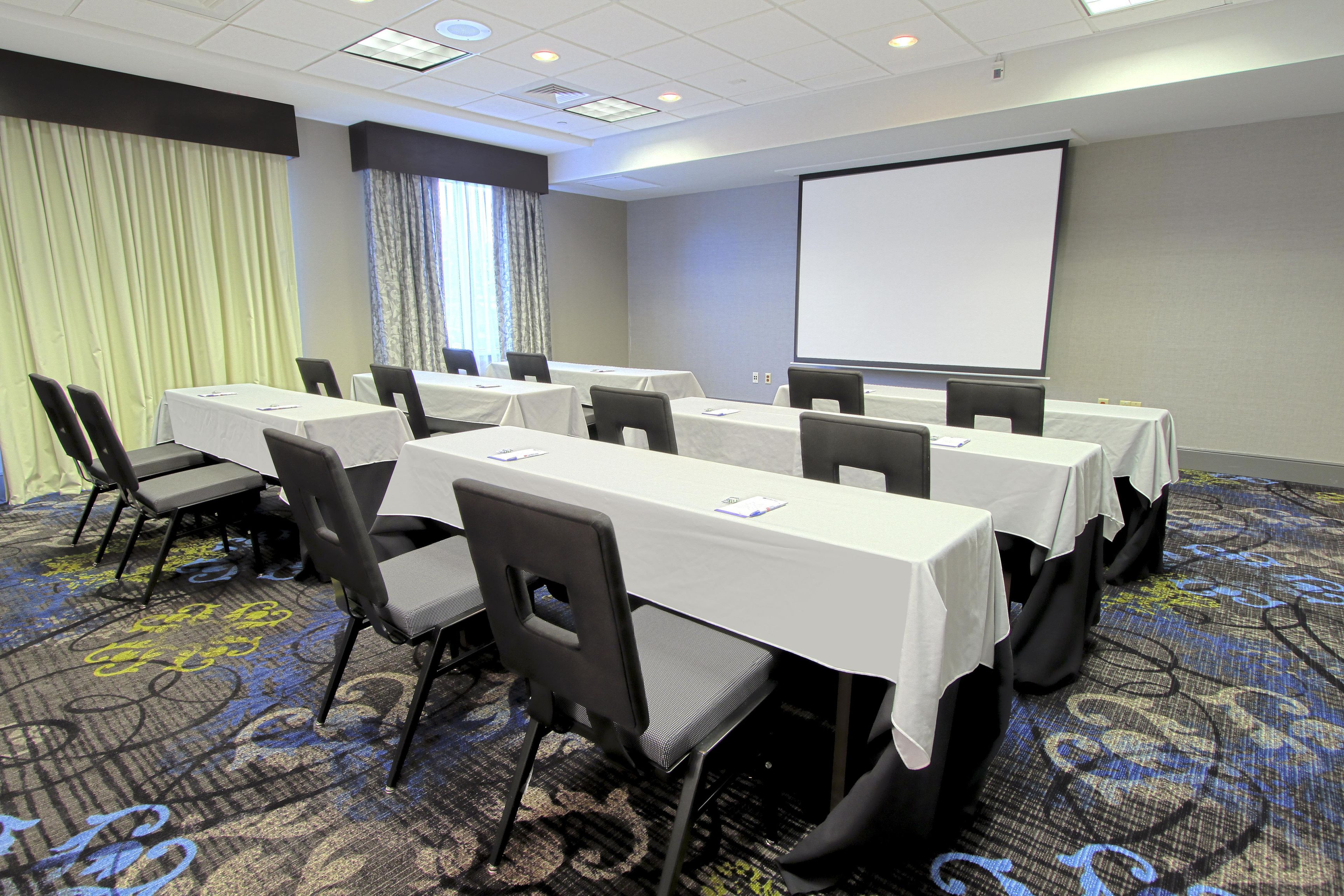A conference room with tables and chairs, a projector screen, and a large window with curtains.