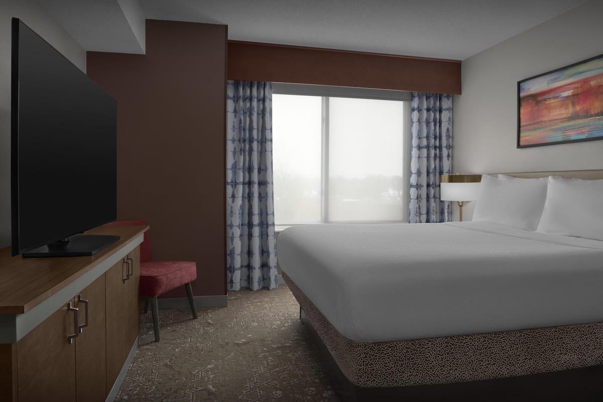 Ett hotellrum med en säng, en tv-apparat på ett träskåp, en röd stol och ett stort fönster med gardiner.