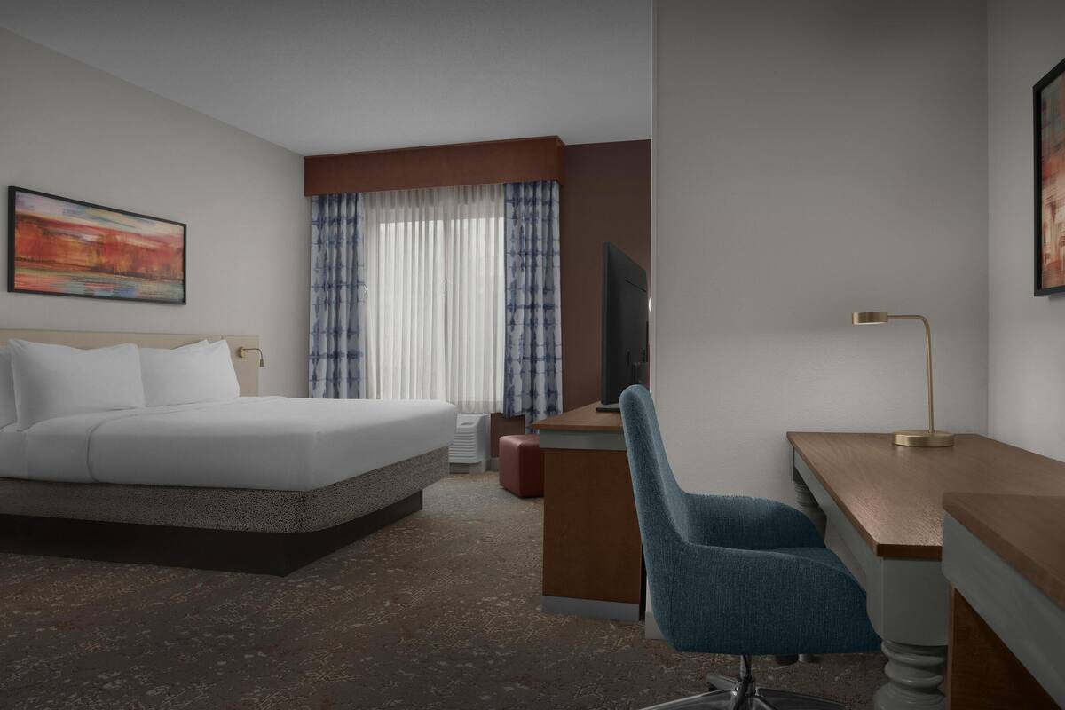 Ett hotellrum med en säng, ett skrivbord, en stol, en TV och gardiner.