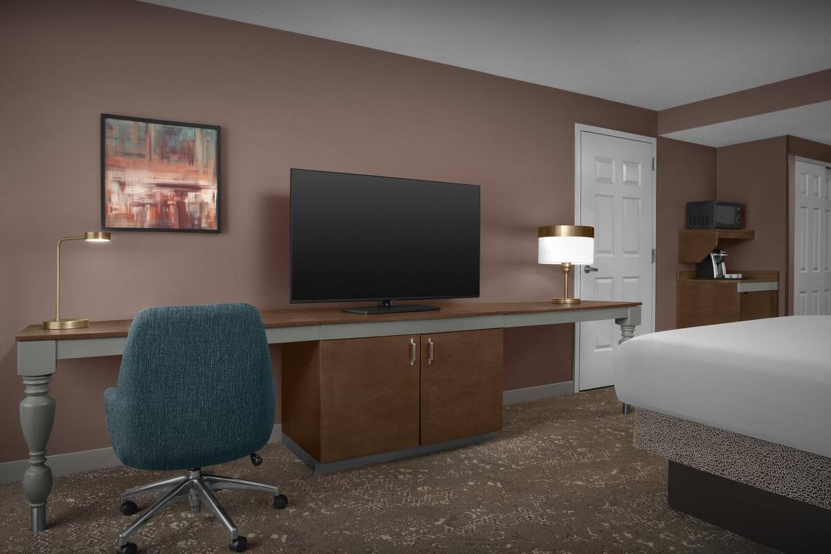 Ett hotellrum med en säng, ett skrivbord med en tv, en stol, en lampa och en tavla på väggen.