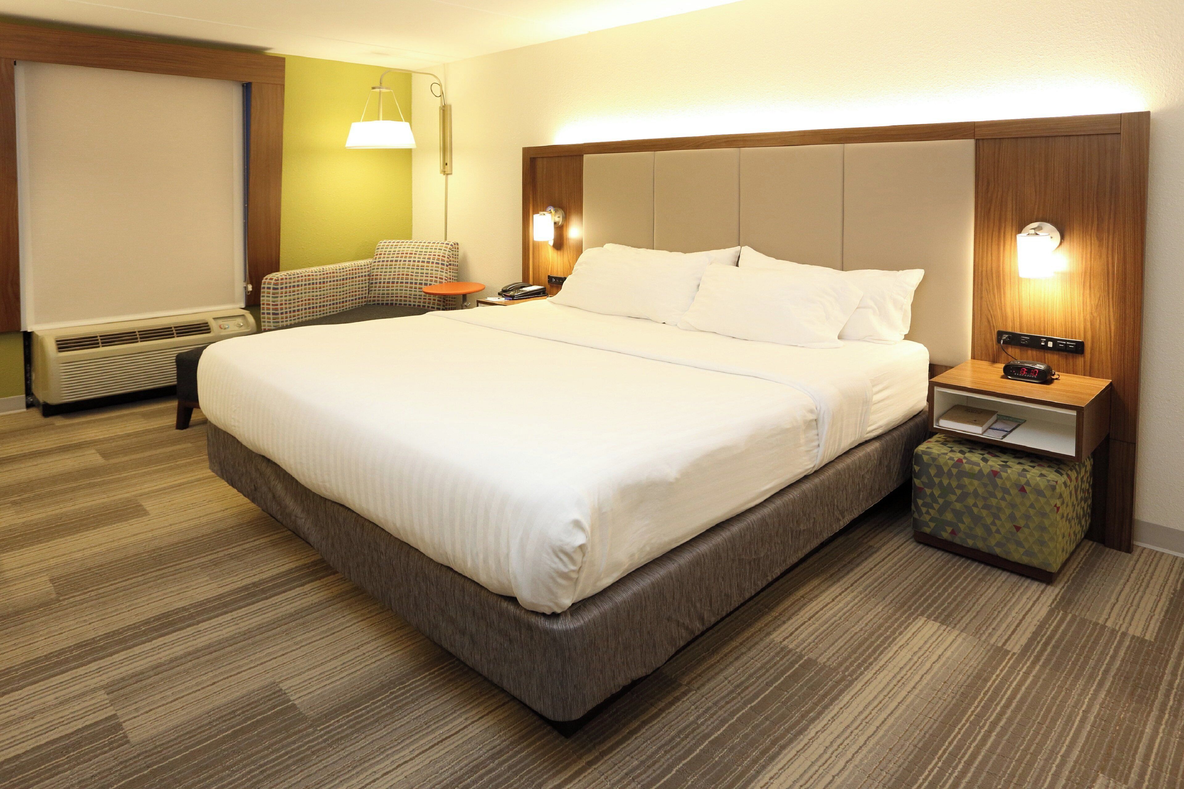 Una habitación de hotel con una cama grande, una mesita de noche con lámpara, una silla y un pequeño reposapiés.