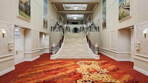 A grand hotel lobby with a marble staircase, ornate carpet, and large landscape paintings on the walls.