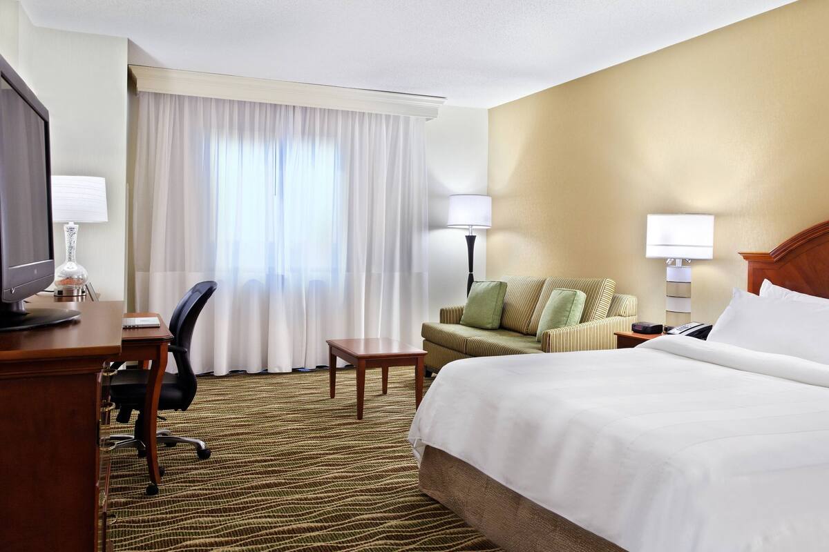 Ein Hotelzimmer mit einem Bett, einem Schreibtisch mit Computer, einem Sessel und einem Fenster mit Vorhängen.