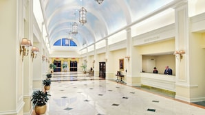 A spacious lobby with a high ceiling, chandeliers, a registration desk, and potted plants.