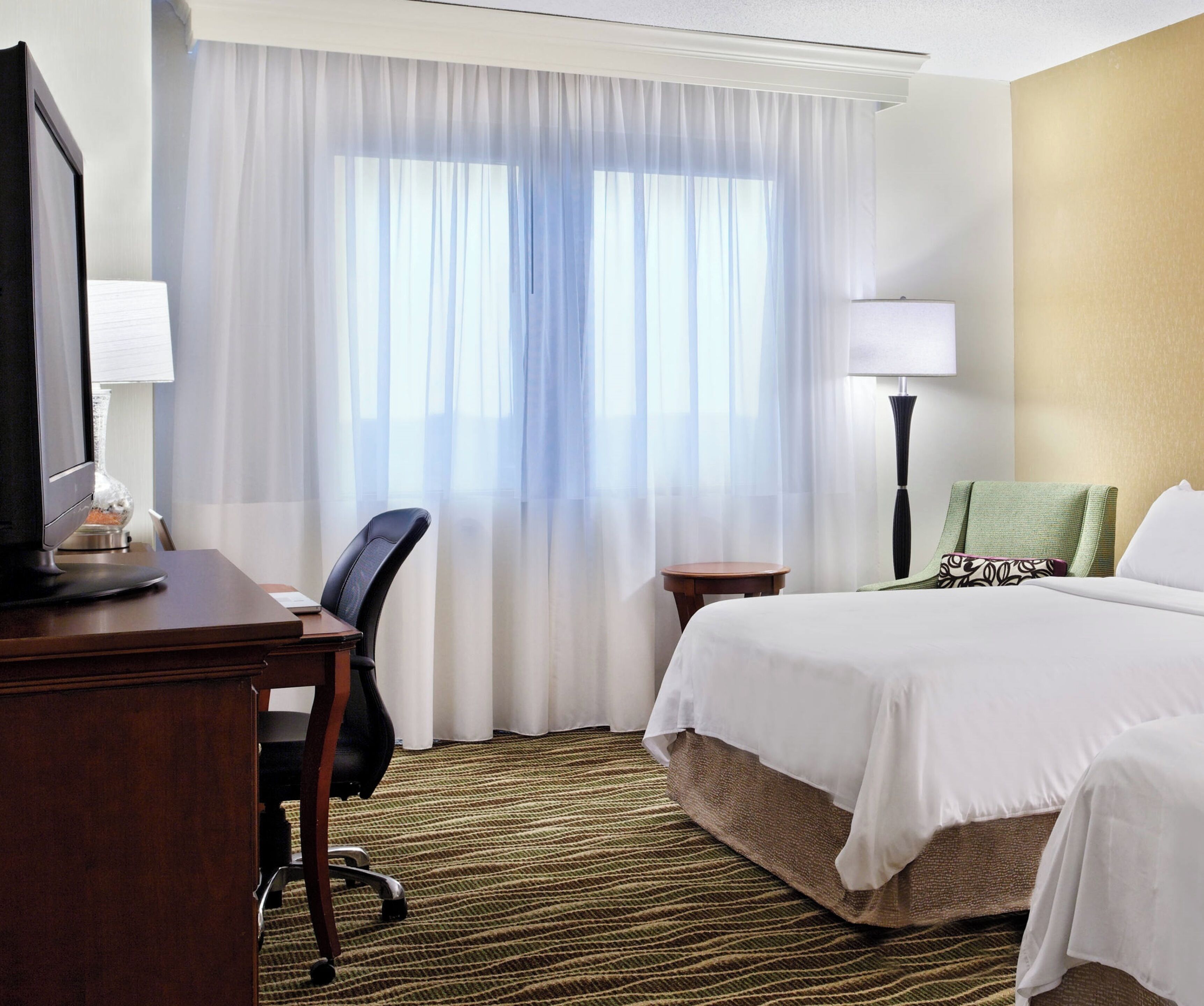 A hotel room with a bed, a desk with a computer, a chair, a lamp, and a window with curtains.