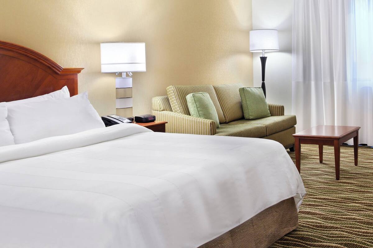 Ein Hotelzimmer mit einem großen Bett, einem gestreiften Sessel, einem kleinen Tisch, einer Lampe und einem Nachttisch mit Telefon und Fernbedienung.