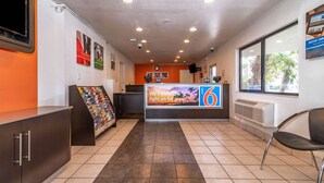 A reception area with a counter, a clock, and a window with a view of palm trees. - Motel 6 Palm Springs, CA - East - Palm Canyon (Palm Springs)