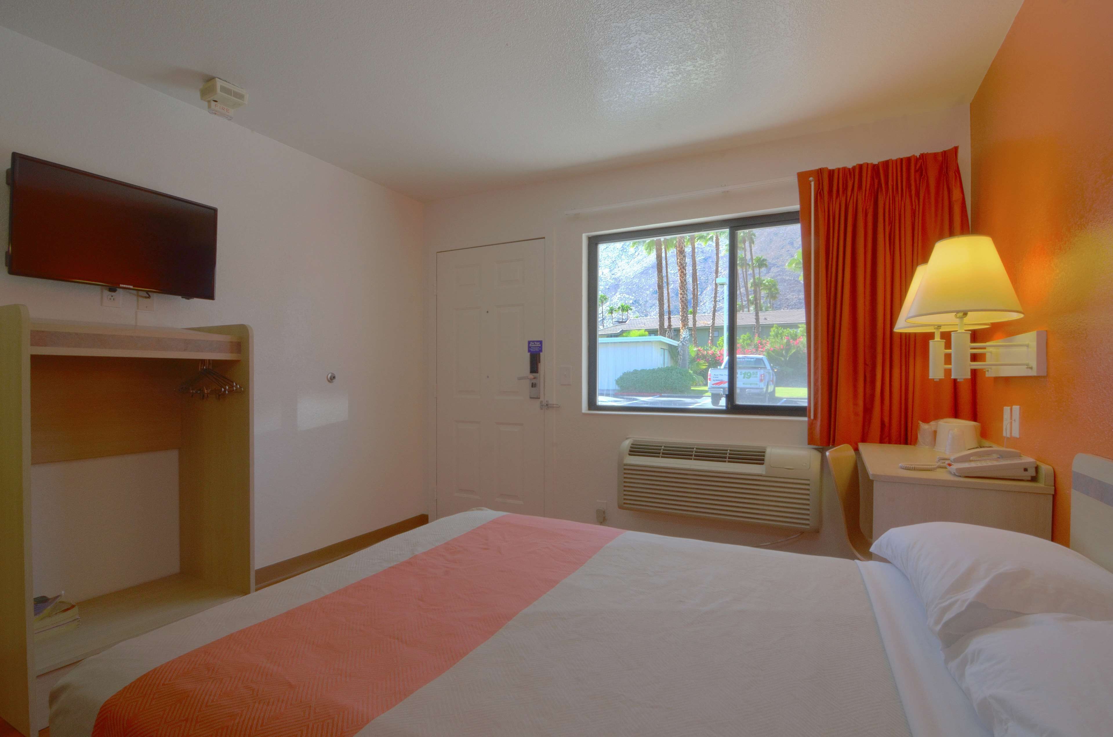 A hotel room with a bed, a TV, a window with curtains, a desk with a lamp, and a heater.
