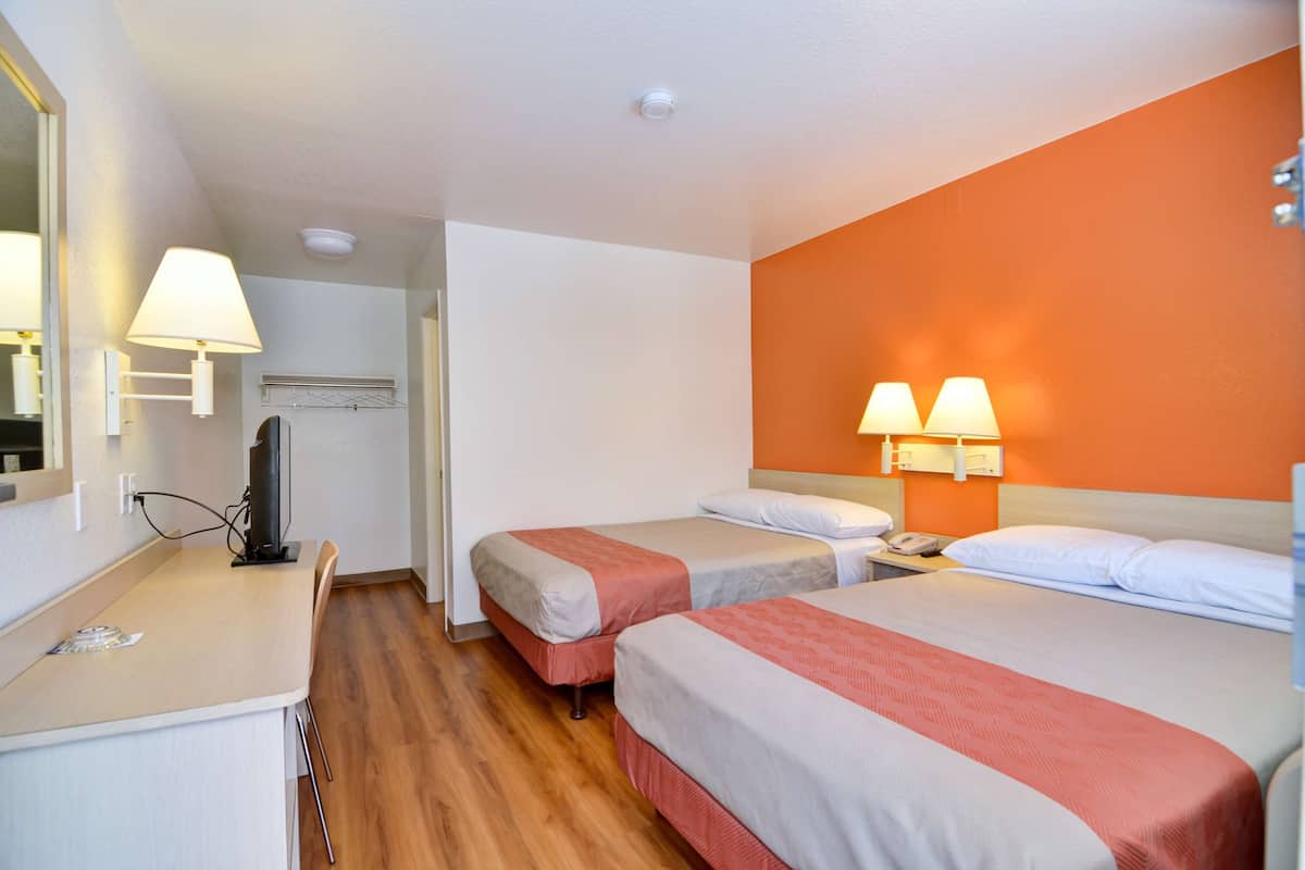 A hotel room with two beds, a desk, a mirror, and a wall-mounted lamp.