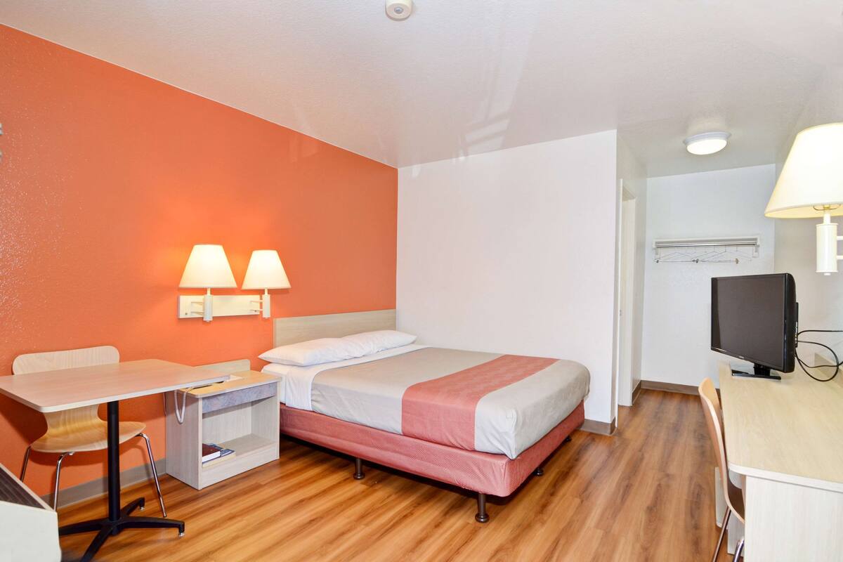 A compact hotel room with a bed, a desk, a chair, a TV, and a lamp.